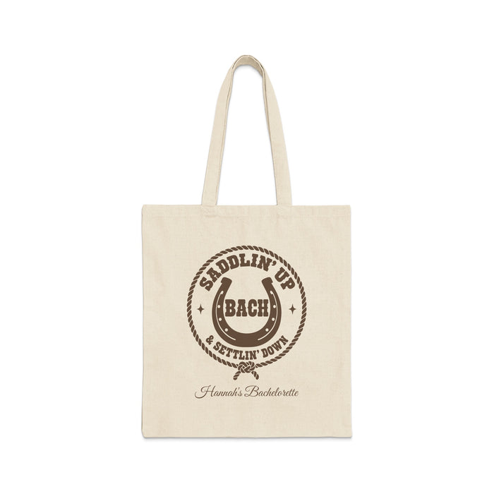 Off-white canvas tote bag with brown rodeo-themed text, horseshoe, and rope graphic on the front.