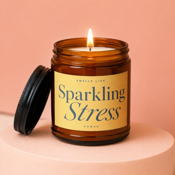 Amber jar candle burning with a black lid to the side, labeled "Sparkling Stress VOWED".