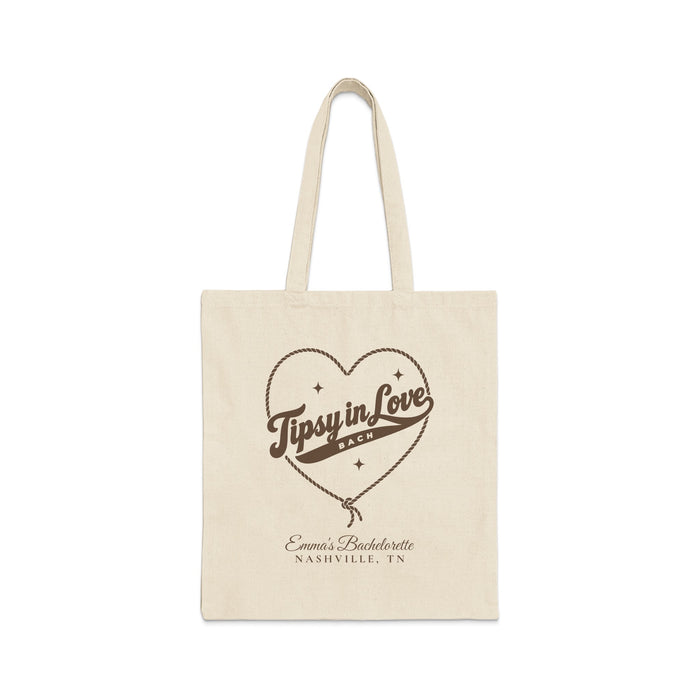 A plain canvas tote bag featuring brown print with a rope-outlined heart, stars, and text reading Tipsy in Love Bach.