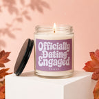 Candle with "Officially Dating Engaged Vowed" label on pink background, surrounded by leaves.