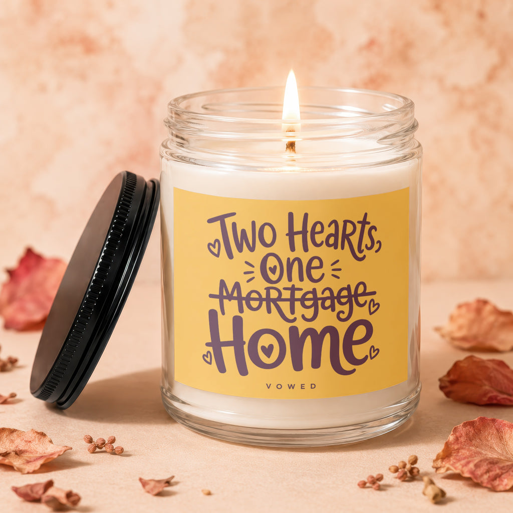 Lit glass jar candle with yellow label reading "Two Hearts, One Mortgage Home".