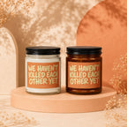 Two candles with black lids and orange labels that read "WE HAVEN'T KILLED EACH OTHER YET".