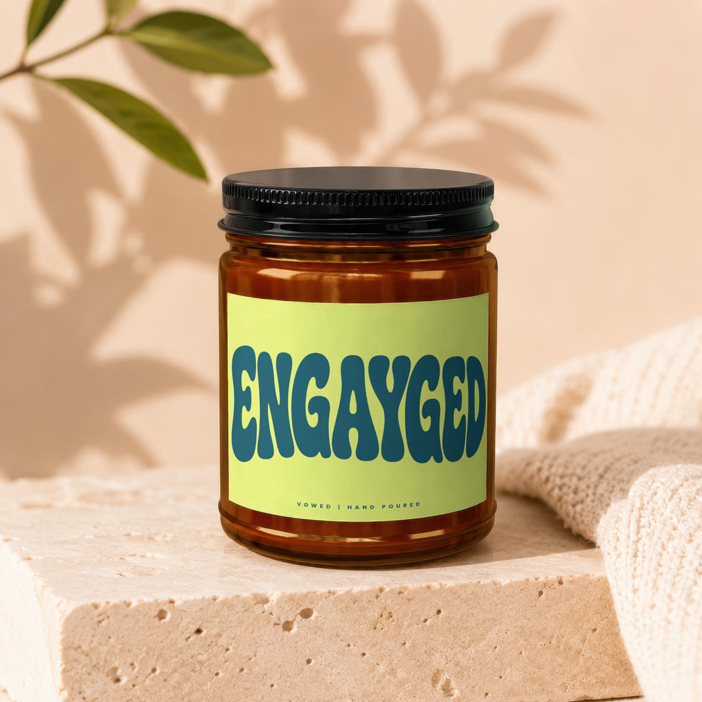 Amber jar candle with "ENGAYGED" text on a lime green label on a stone block.