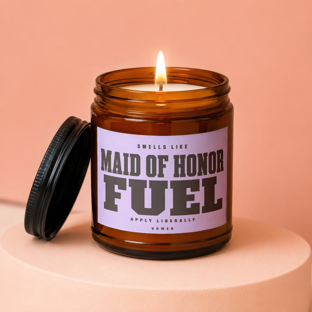 A lit amber jar candle with "MAID OF HONOR FUEL" text, lid ajar.