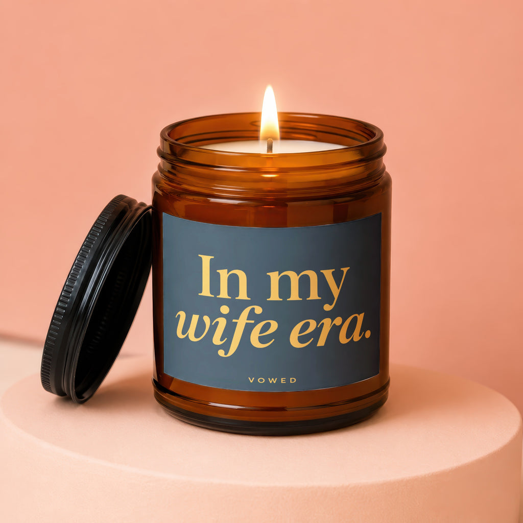 Amber jar candle with lit wick and black lid that reads "In my wife era. VOWED."