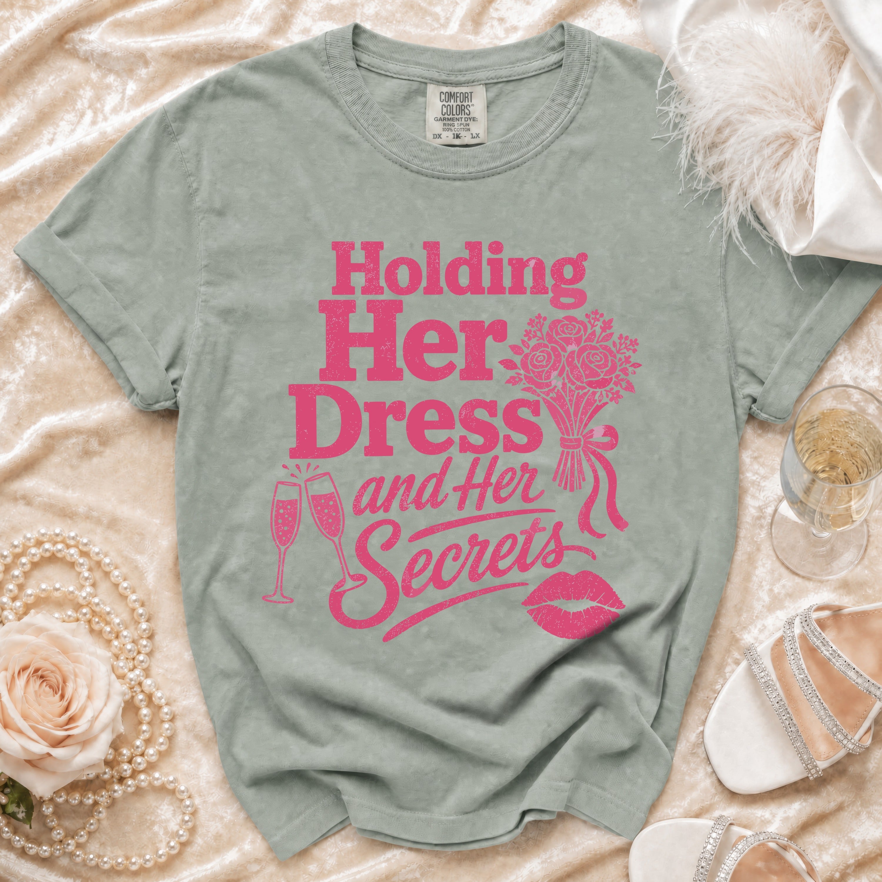 T-shirt with pink text "Holding Her Dress and Her Secrets", champagne glasses, lips, and bouquet.