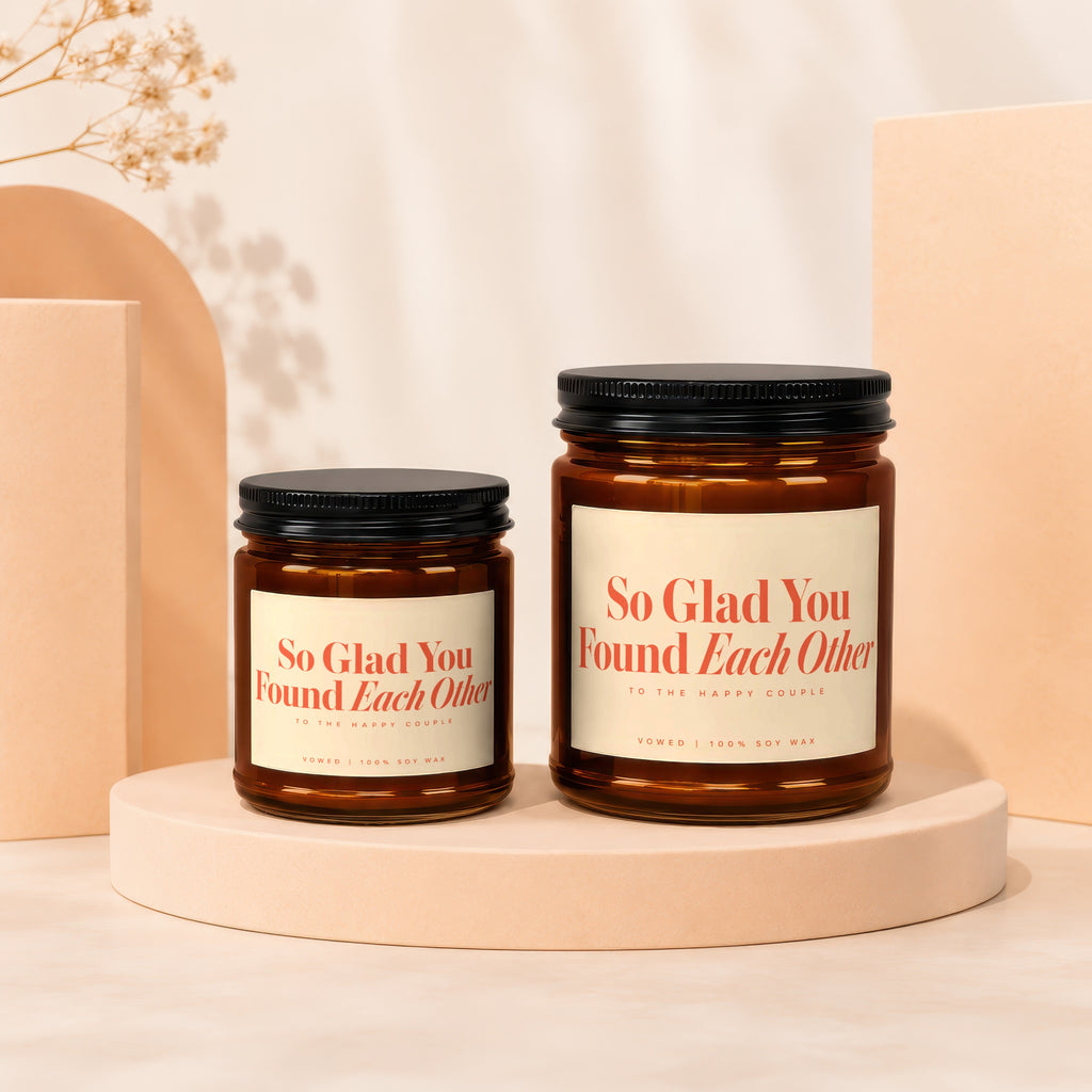 Two amber glass candles with black lids reading "So Glad You Found Each Other."