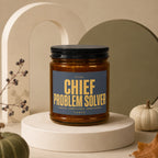 Amber jar candle with "Chief Problem Solver" text, featuring decorative pumpkins and foliage.