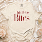 Cream t-shirt with "This Bride Bites" text in red, featuring pearl necklace and bridal shoes.