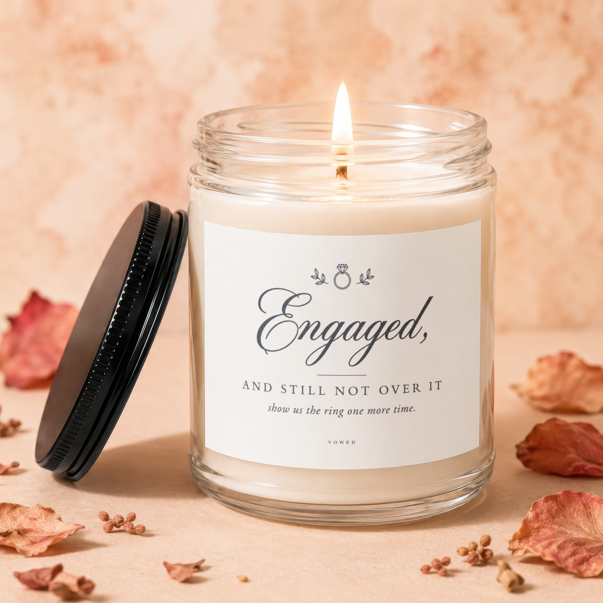 Glass jar candle with lit wick and label reading "Engaged, and still not over it".