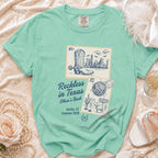 T-shirt with vintage stamps showing a cityscape and cowboy boots, reads "Reckless in Texas Olivia's Bach".