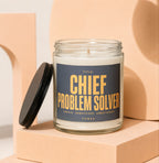 Candle with "Chief Problem Solver" text on dark label and black lid askew.