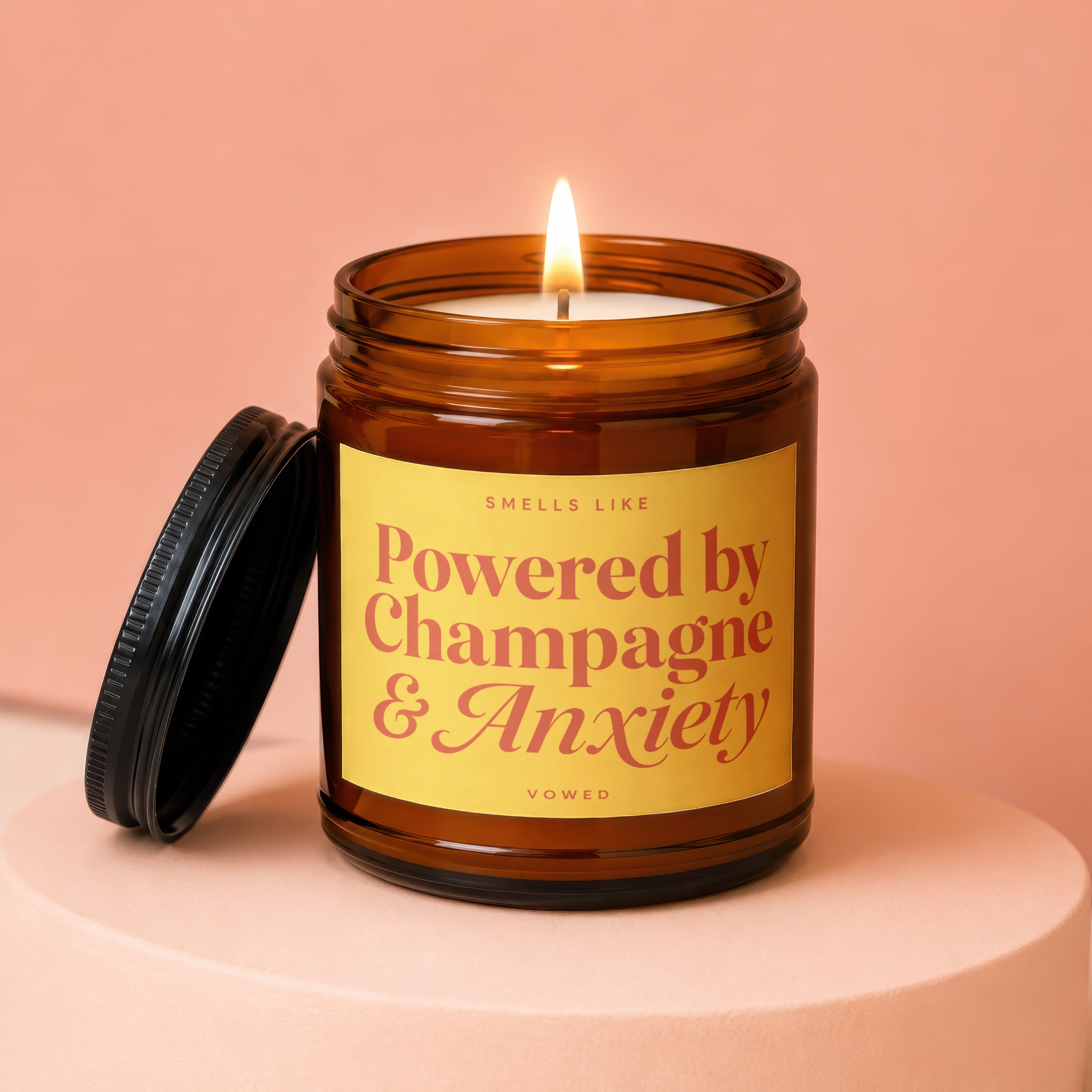 Amber candle with lit wick, yellow label reading "Smells Like Powered by Champagne & Anxiety".