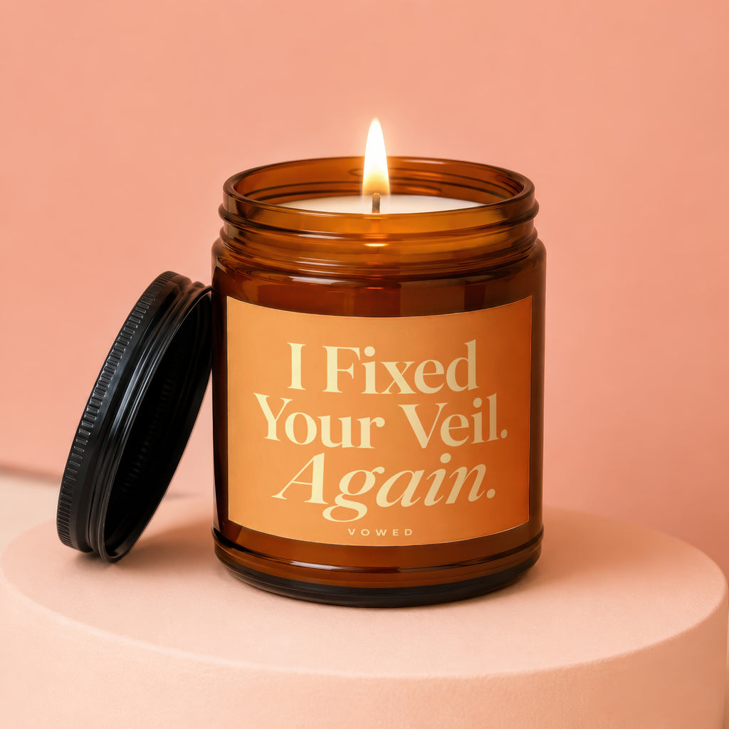 Lit amber candle in glass jar with orange label reading "I Fixed Your Veil. Again."