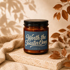 Amber jar candle with black lid, "Worth the Toaster Oven" text, on a stone block.
