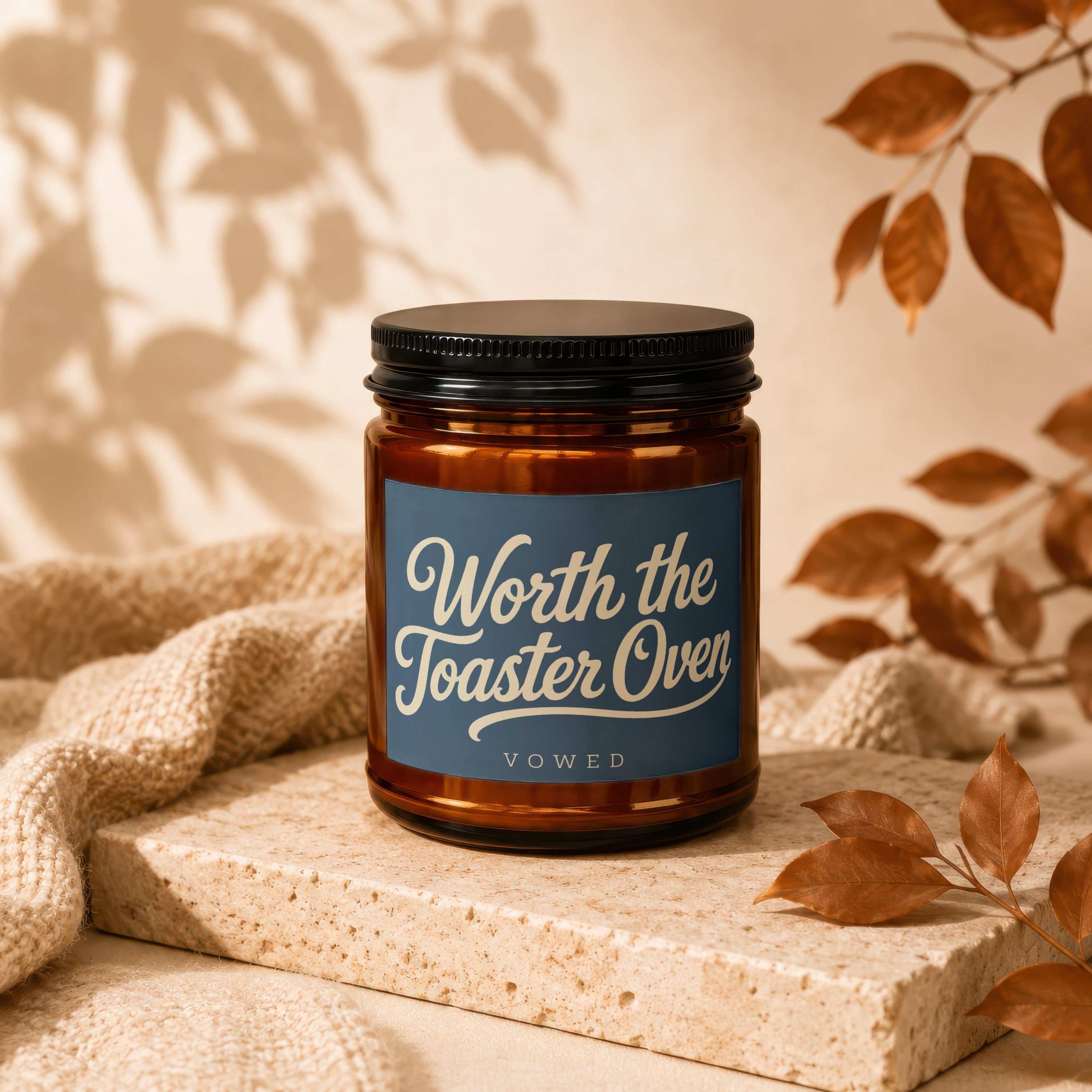 Amber jar candle with black lid, "Worth the Toaster Oven" text, on a stone block.