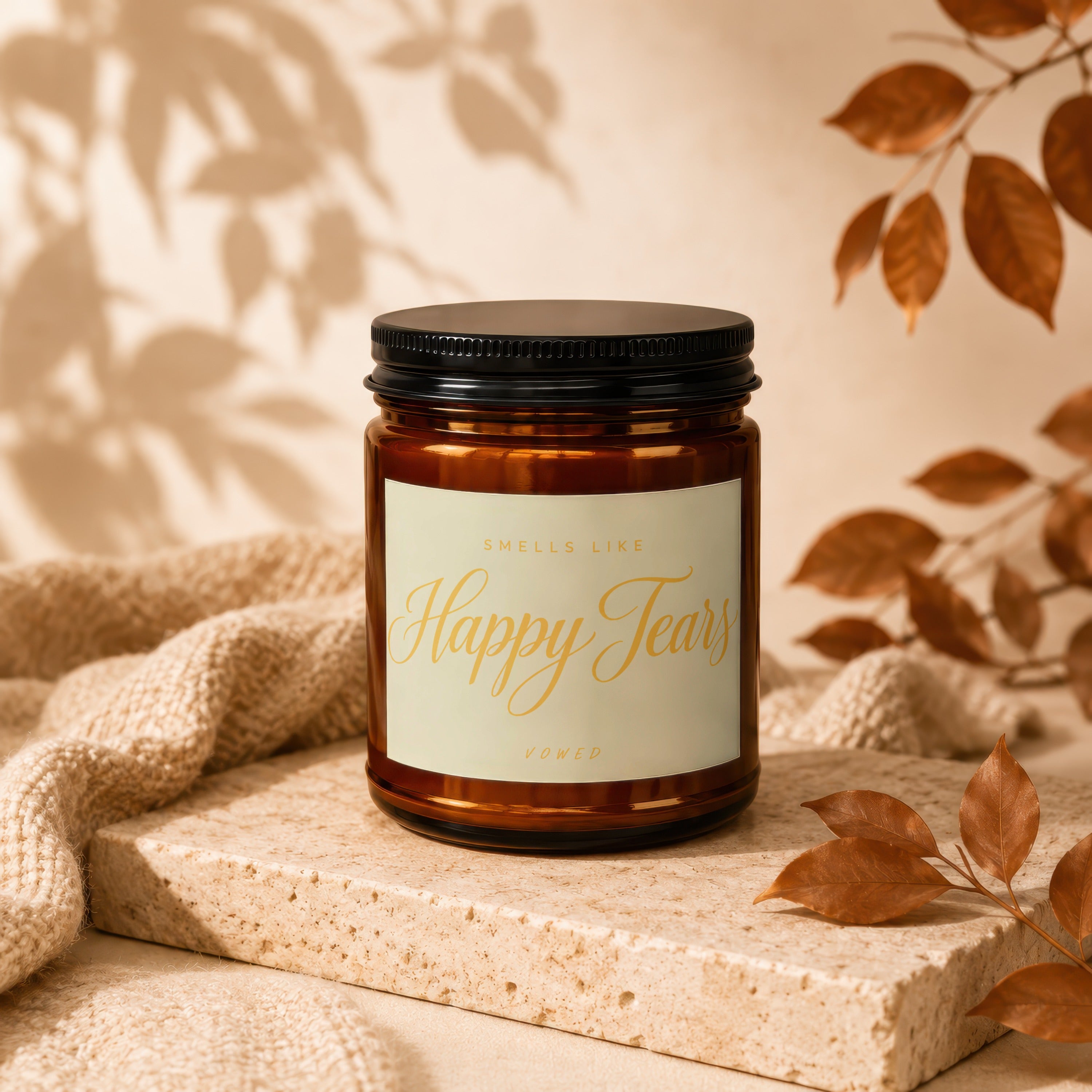 Brown glass candle with "Smells Like Happy Tears" label, on textured stone and fabric with autumn leaves.