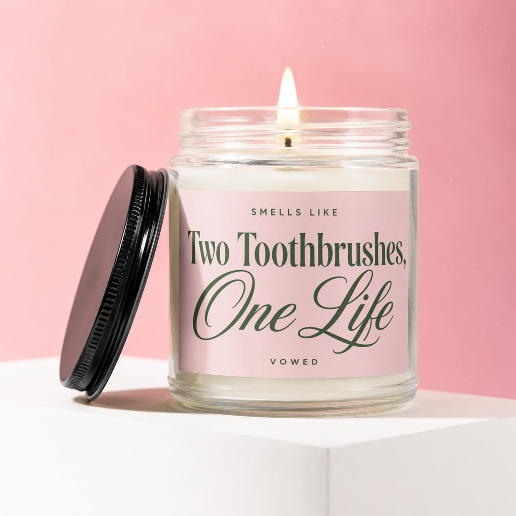 Jar candle with lit wick, black lid resting beside it, and pink label reading "Smells Like Two Toothbrushes, One Life Vowed".