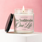 Jar candle with lit wick, black lid resting beside it, and pink label reading "Smells Like Two Toothbrushes, One Life Vowed".