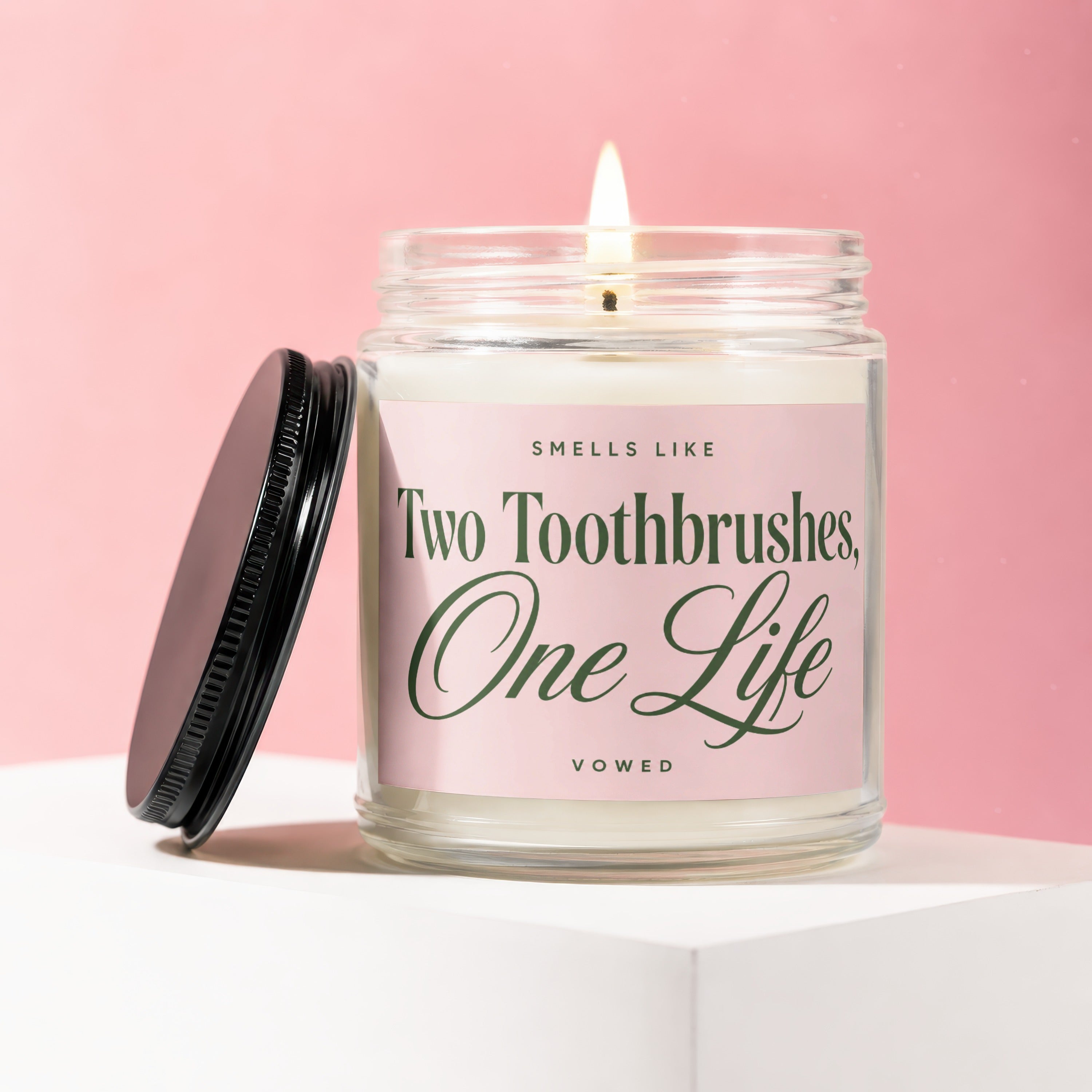 Jar candle with lit wick, black lid resting beside it, and pink label reading "Smells Like Two Toothbrushes, One Life Vowed".
