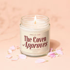 Candle with "The Coven Approves" text on the label, surrounded by petals.