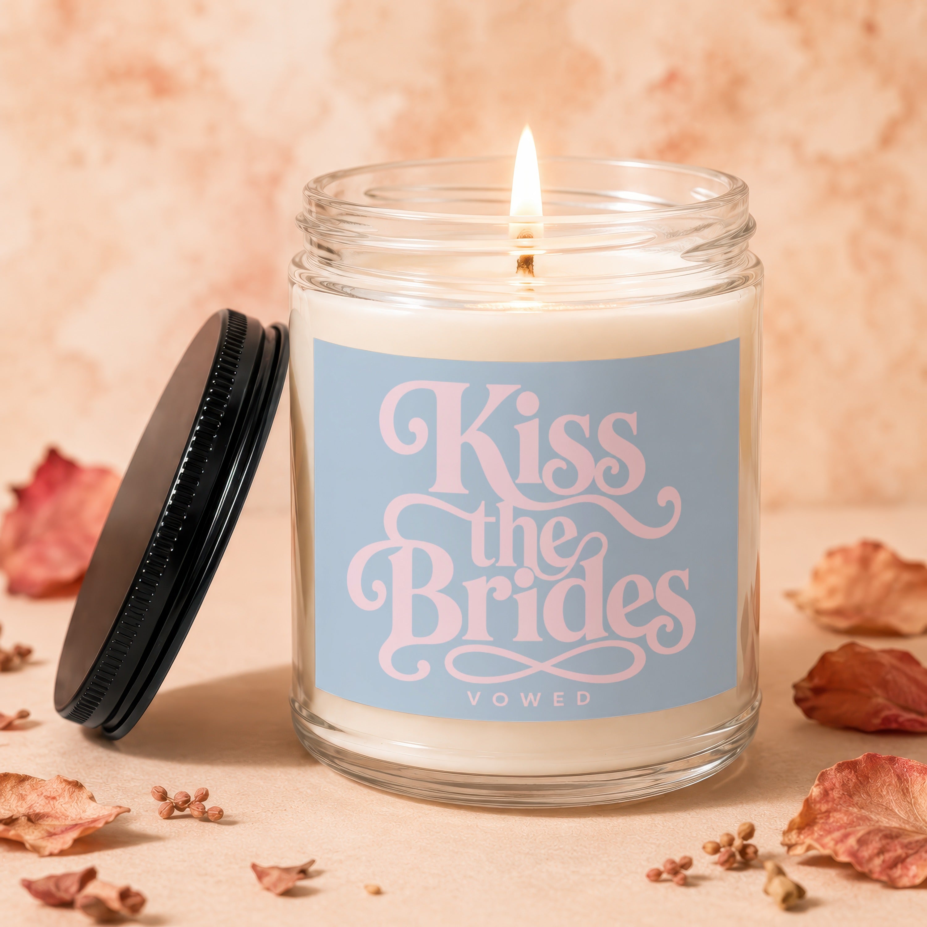 Glass jar candle with a lit wick and a blue label that reads "Kiss the Brides VOWED".