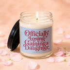 Candle with lid off, lit wick, and pink text: "Officially Losing, Gaining a Daughter."