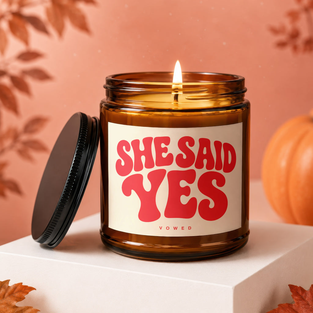 Amber jar candle with lit flame, "SHE SAID YES" text, and "VOWED" subtitle.