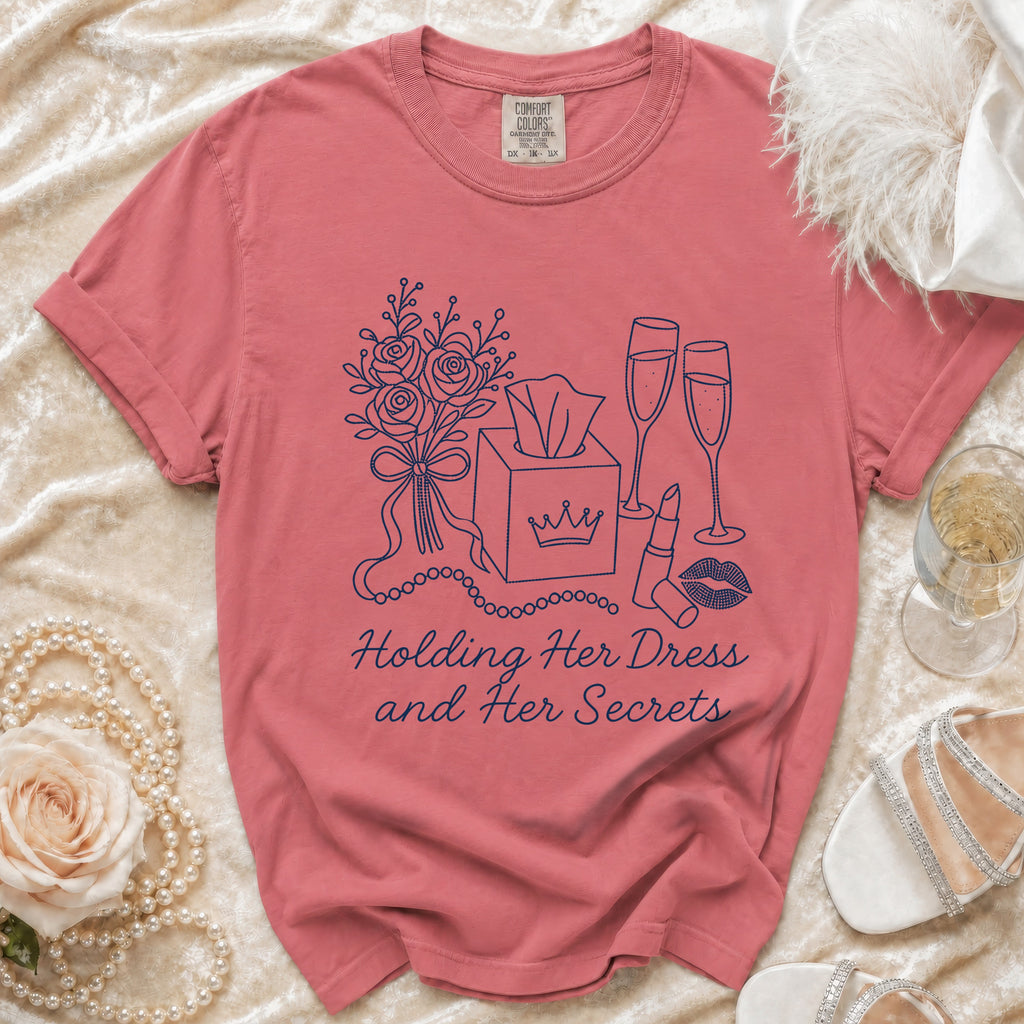 Rose-colored t-shirt with line art of flowers, tissues, champagne, and lipstick. Text reads: Holding Her Dress and Her Secrets.