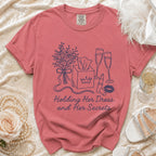 Rose-colored t-shirt with line art of flowers, tissues, champagne, and lipstick. Text reads: Holding Her Dress and Her Secrets.