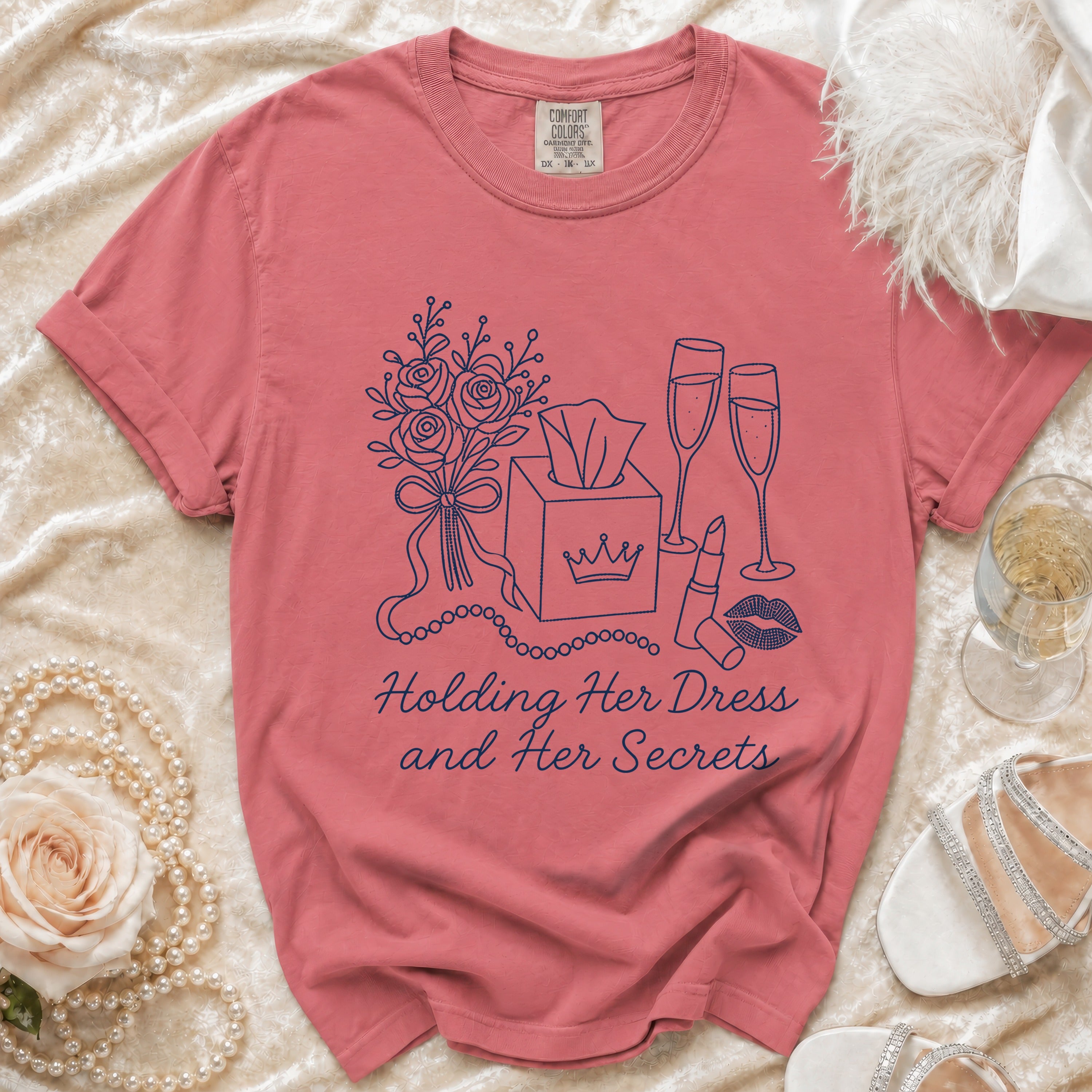 Rose-colored t-shirt with line art of flowers, tissues, champagne, and lipstick. Text reads: Holding Her Dress and Her Secrets.