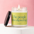 Glass candle with lit wick on a white surface with a black lid and pink background.