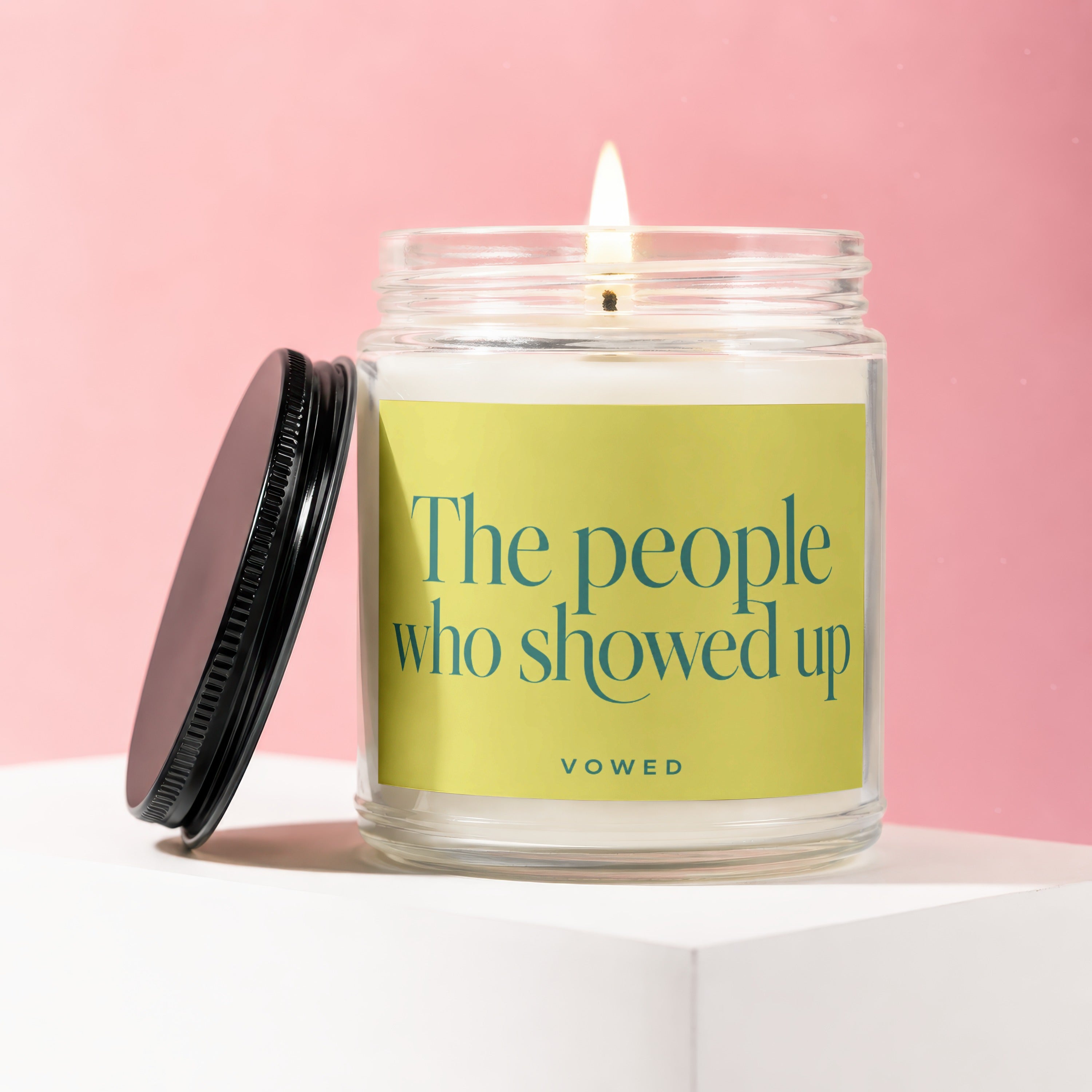 Glass candle with lit wick on a white surface with a black lid and pink background.