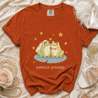 Orange t-shirt with cartoon frogs wearing wedding attire and "Hoppily Hitched" text.