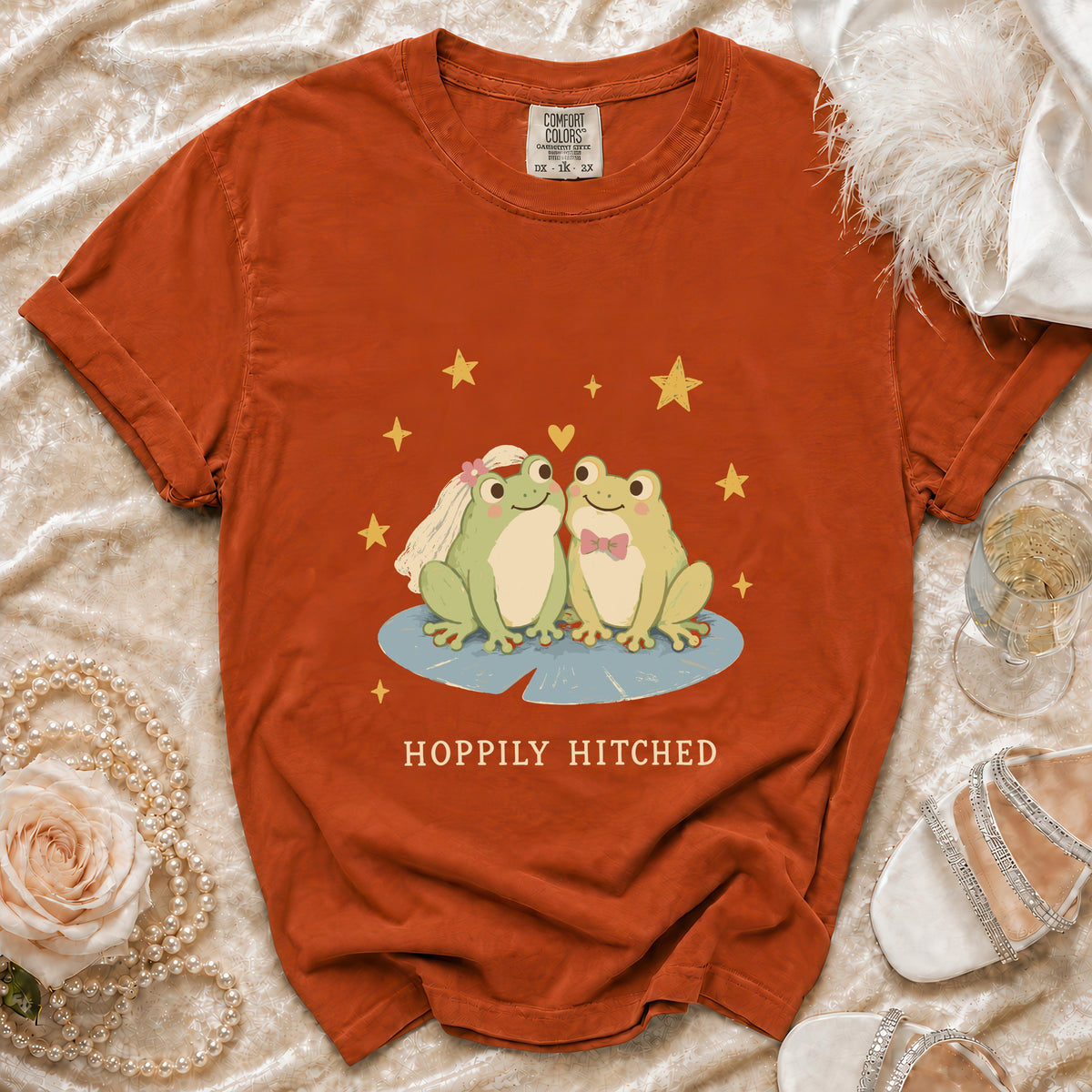 Orange t-shirt with cartoon frogs wearing wedding attire and "Hoppily Hitched" text.