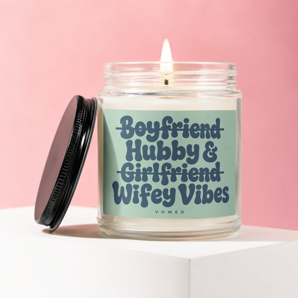Glass candle with lit flame and black lid, says "Boyfriend Hubby & Girlfriend Wifey Vibes".