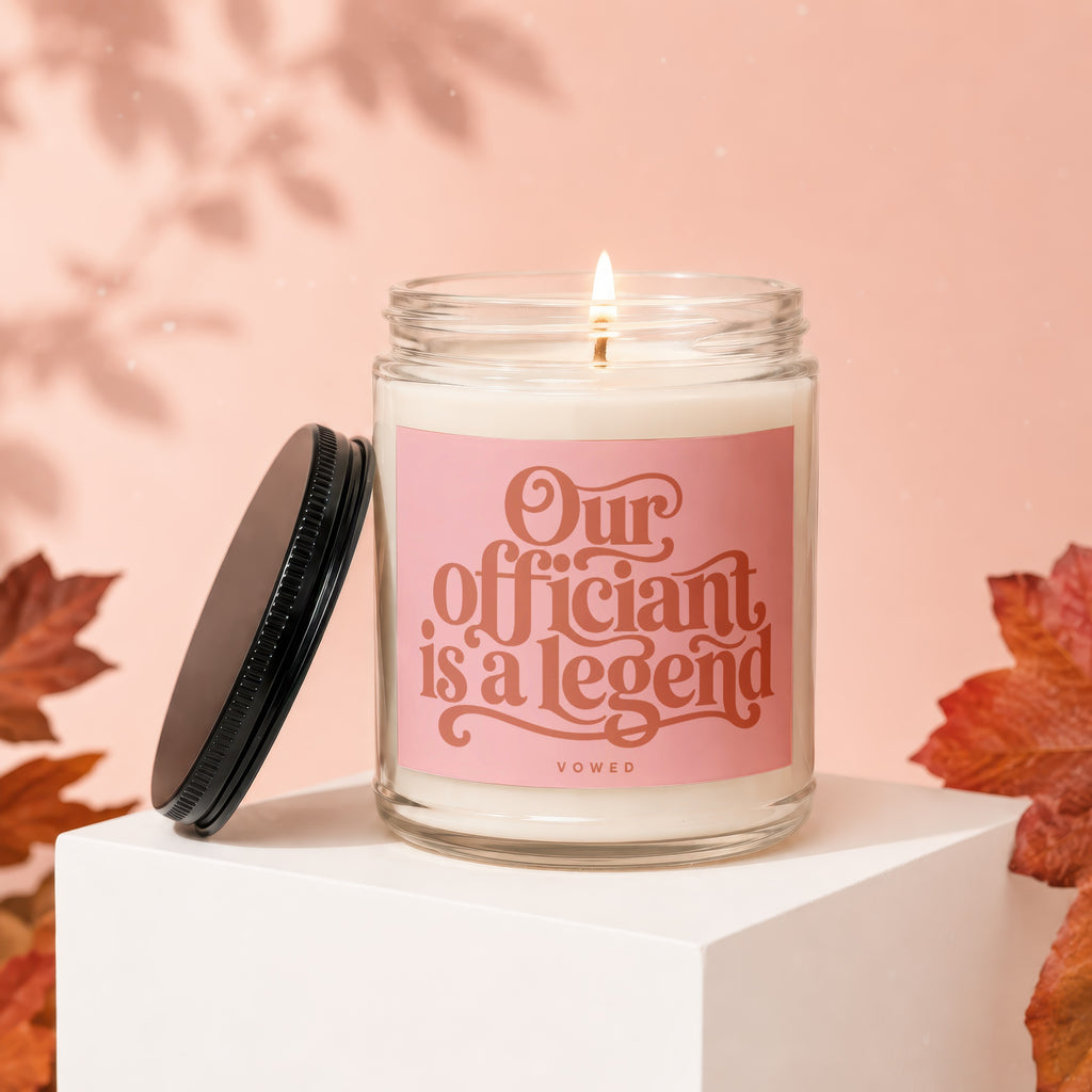 Glass candle with lit wick and pink label reading "Our officiant is a legend VOWED."