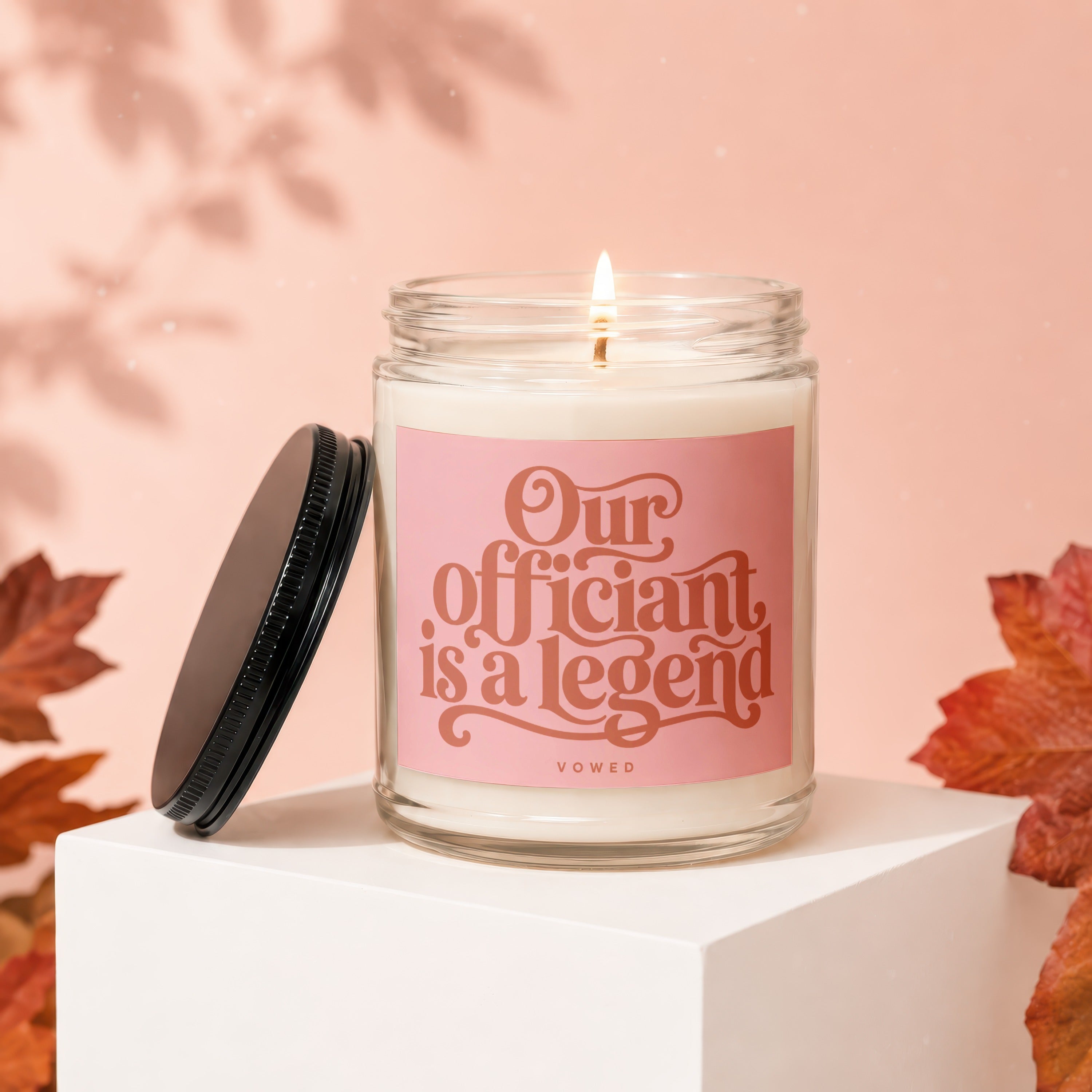 Glass candle with lit wick and pink label reading "Our officiant is a legend VOWED."
