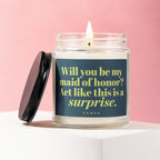 Glass candle with lit wick and dark lid, reading "Will you be my maid of honor? Act like this is a surprise."