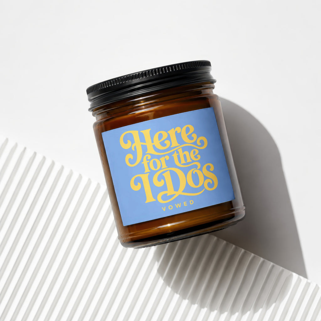 Amber glass candle with black lid, navy label reads "Here for the Dos VOWED".