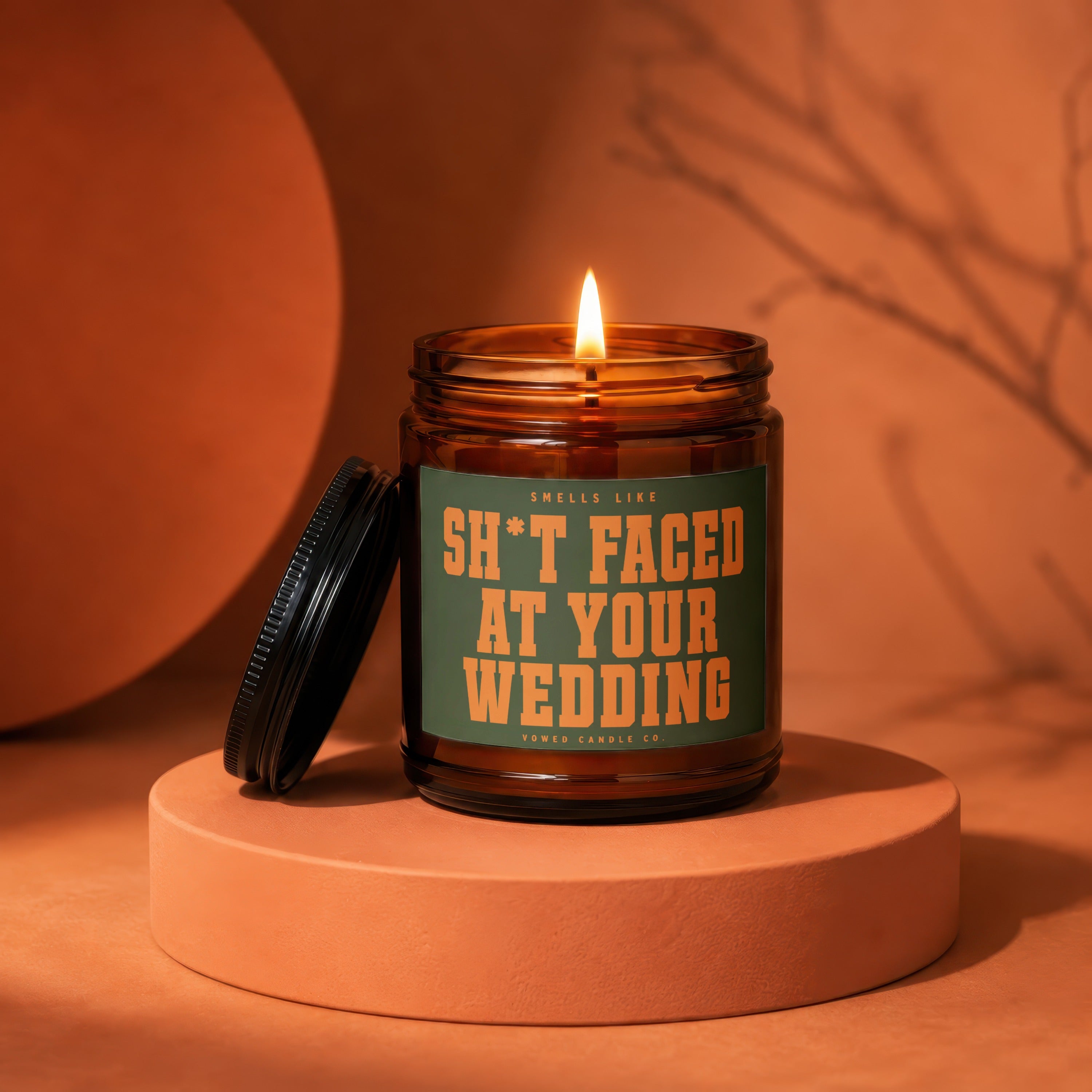 Lit amber candle with label "Smells Like Sh*t Faced At Your Wedding".