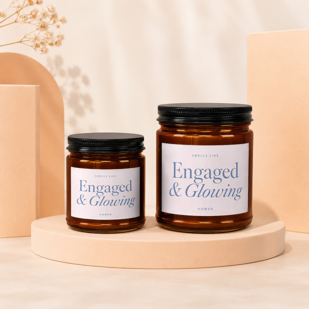 Two amber glass candles with black lids on cream pedestals, labeled "Engaged & Glowing."