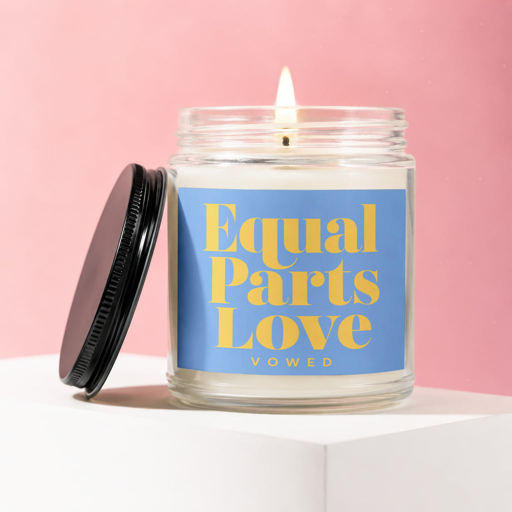 Lit candle in a glass jar with a blue label that reads "Equal Parts Love VOWED"