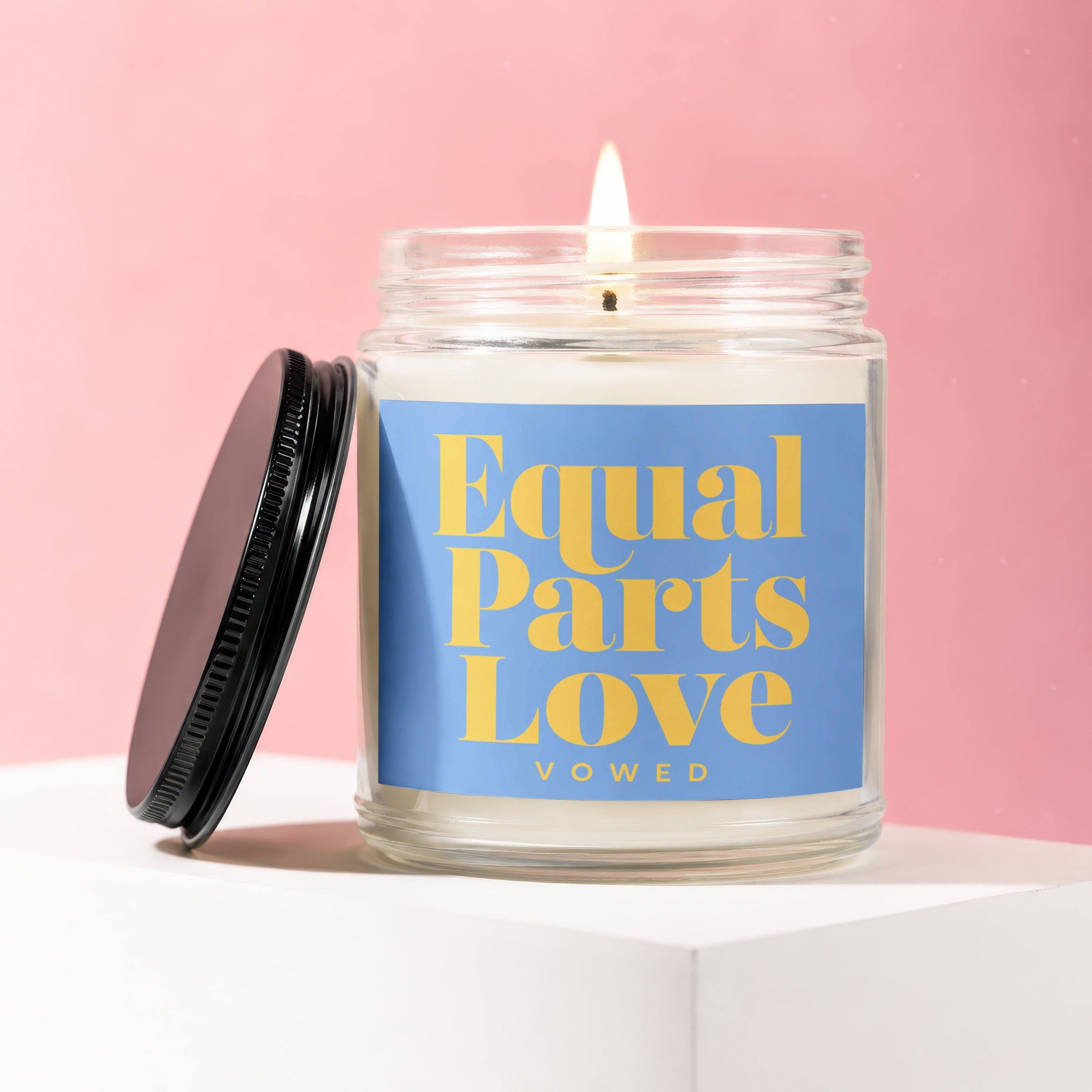 Lit candle in a glass jar with a blue label that reads "Equal Parts Love VOWED"