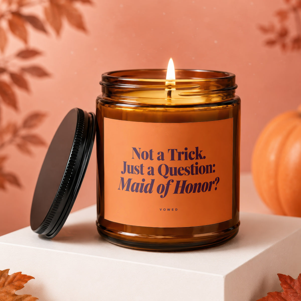 Amber candle with lit flame and orange label reading "Not a Trick. Just a Question: Maid of Honor?".