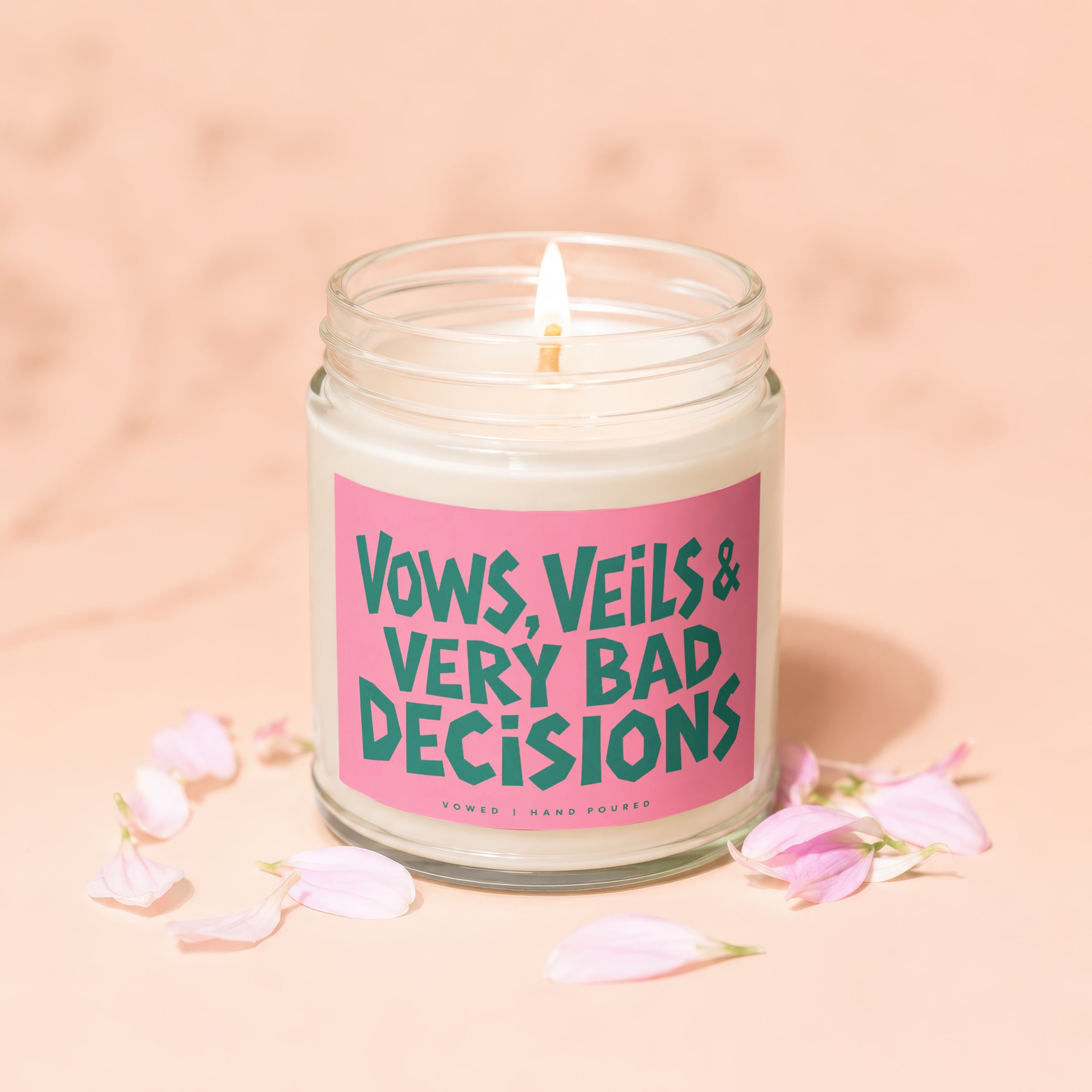 Candle with pink label reading "VOWS, VEILS & VERY BAD DECISIONS" surrounded by petals.