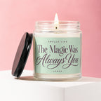 Glass candle with burning wick, lid off, on white pedestal, reads "Smells Like The Magic Was Always You"