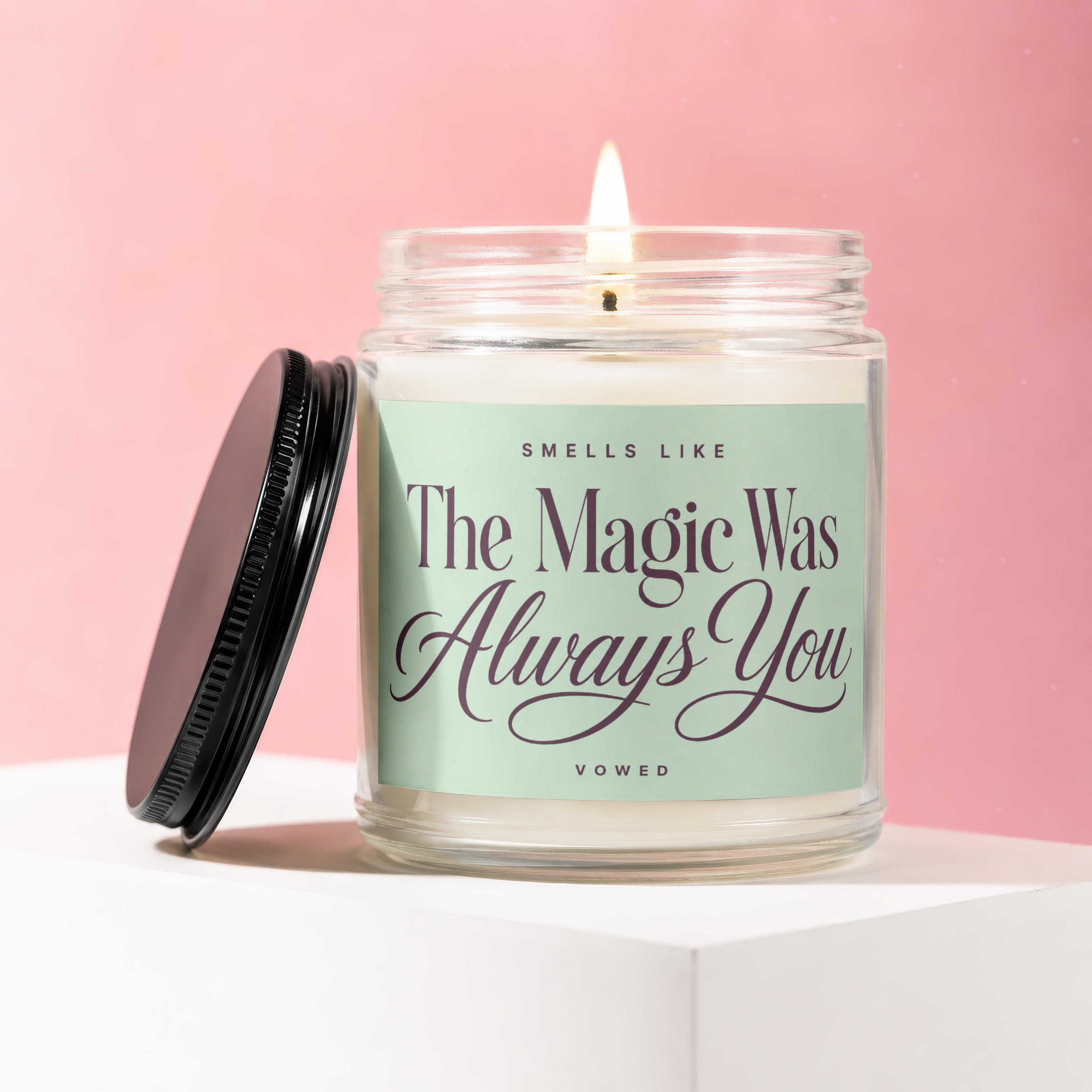 Glass candle with burning wick, lid off, on white pedestal, reads "Smells Like The Magic Was Always You"
