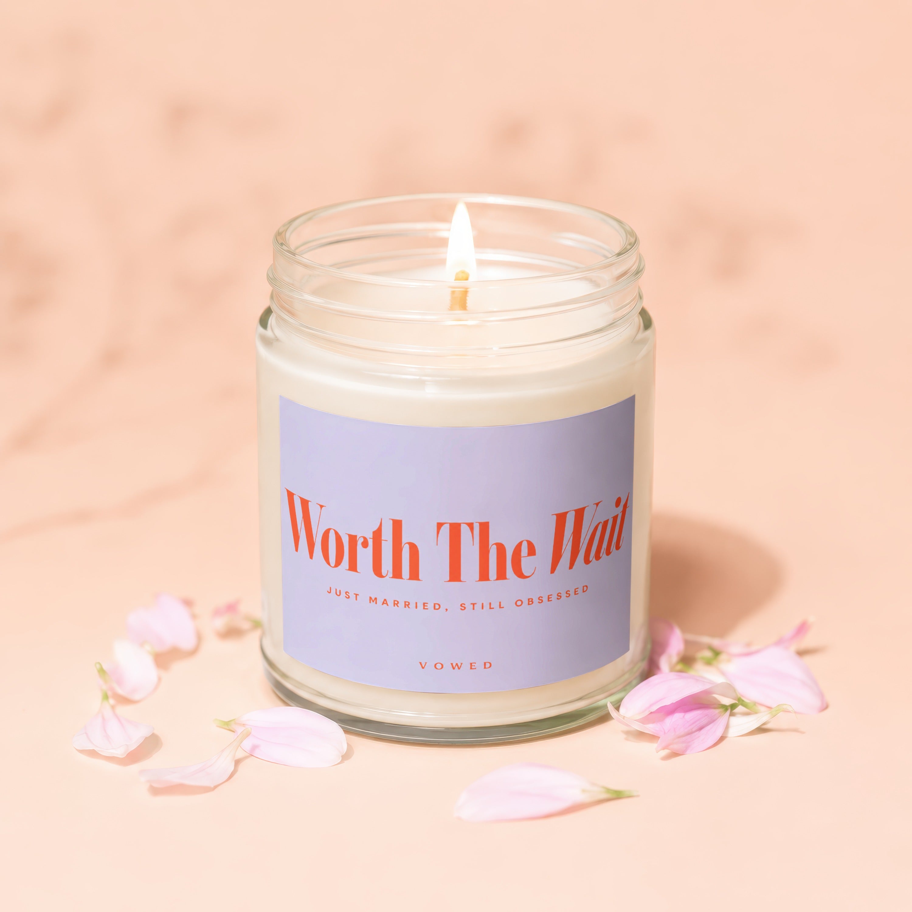 Lit candle in a glass jar with "Worth The Wait" and "Vowed" text, surrounded by petals.
