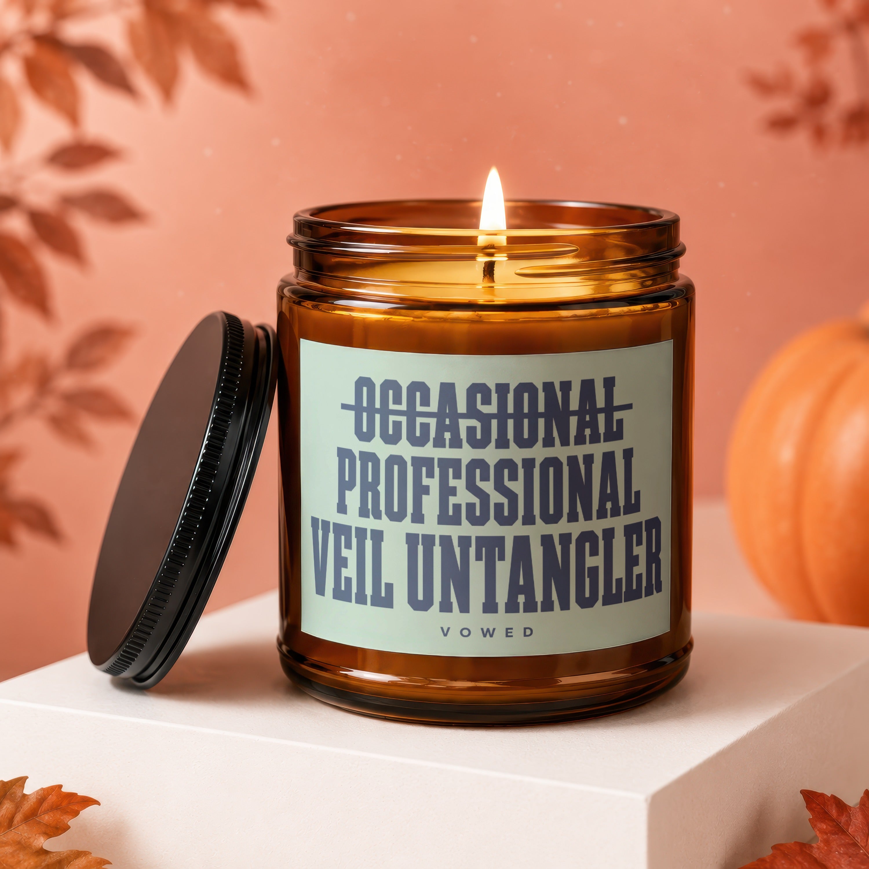 Candle with label "Occasional Professional Veil Untangler Vowed" and lid off, with pumpkin and leaves.